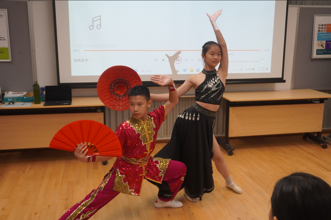 Two student performers in traditional Chinese dance costumes posing with red fans in a classroom.